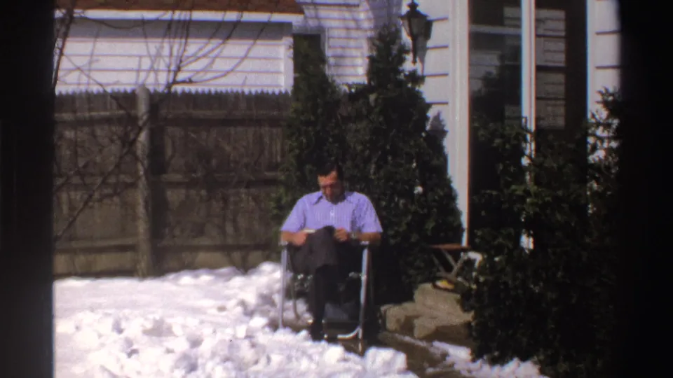 The man remains seated and reading in the snowy Michigan backyard, framed by a wood fence, shrubs, house siding, and a glass door.