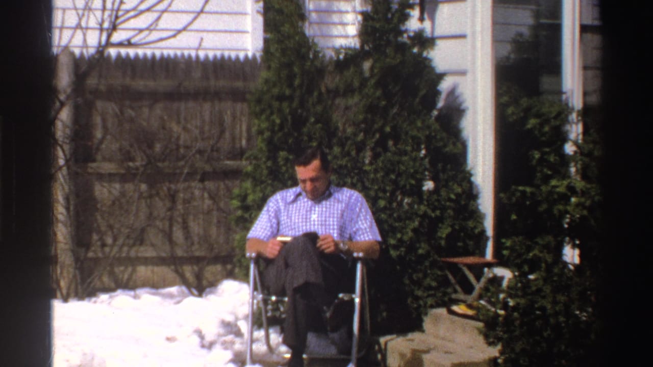A closer 1969 Michigan frame shows the man in a blue and white checkered shirt reading in a folding chair near snow and evergreen shrubs.