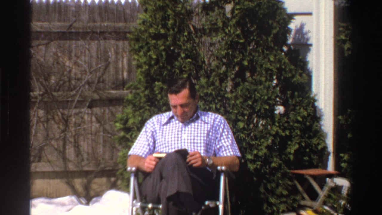 The final frame holds on the man reading in the snowy Michigan yard, with evergreen shrubs, a wood fence, house siding, and an outdoor table behind him.