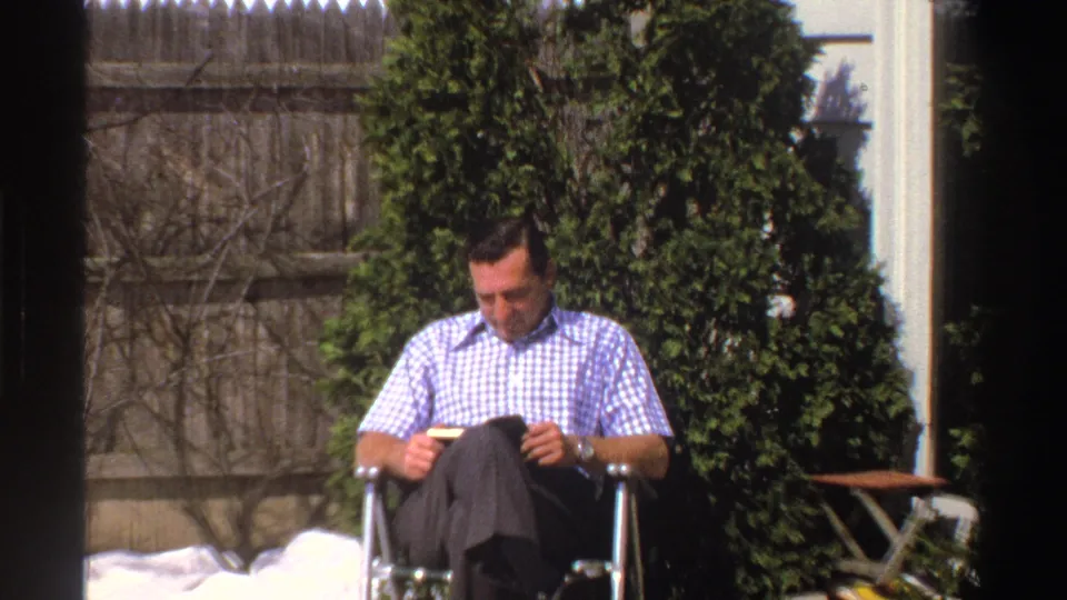 The final frame holds on the man reading in the snowy Michigan yard, with evergreen shrubs, a wood fence, house siding, and an outdoor table behind him.