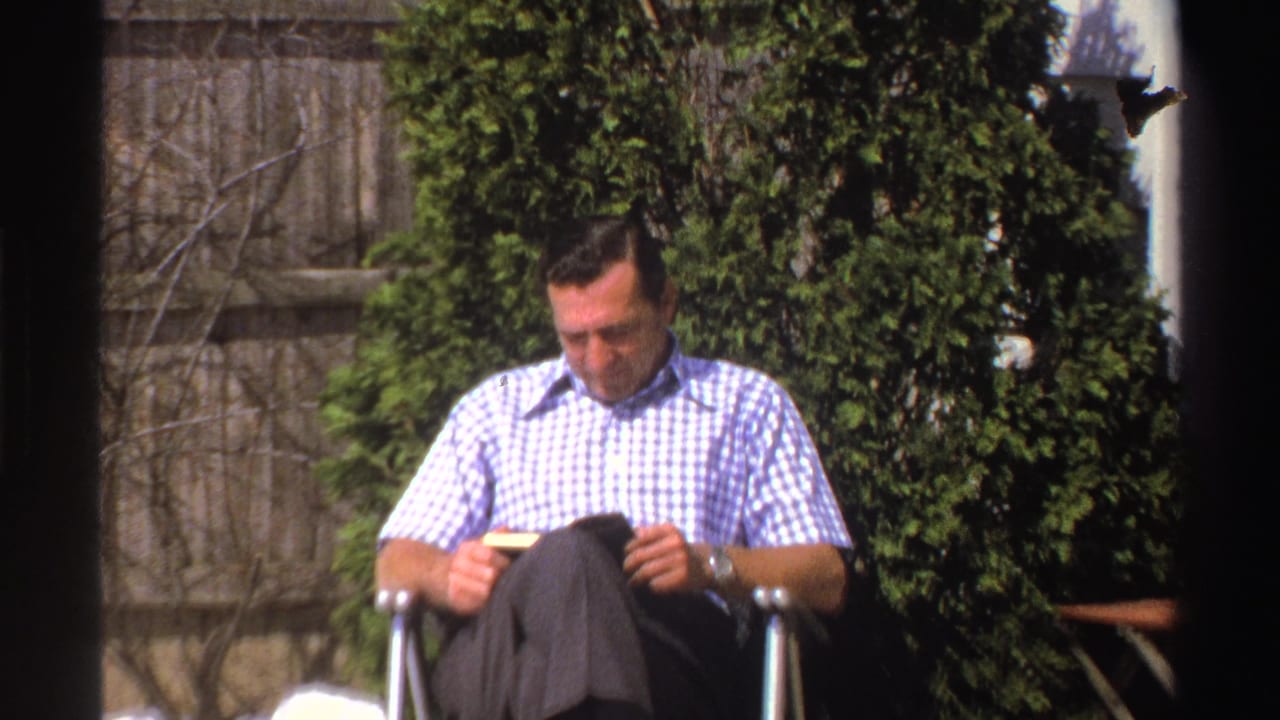 Man in a checkered shirt sits in a backyard chair reading a small book.