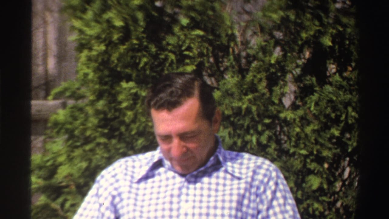 A soft 8mm frame shows the man looking down in a Michigan yard, with shrubs and fencing visible behind him.