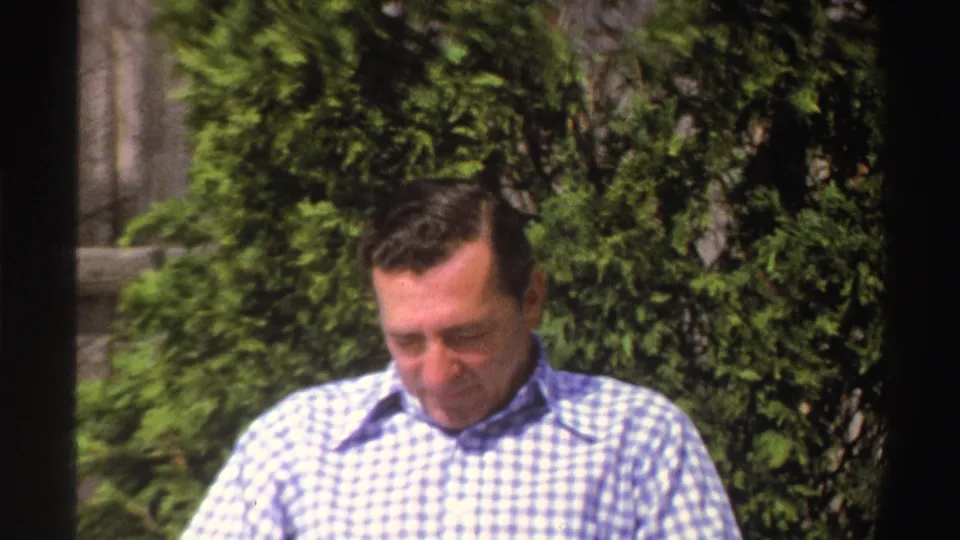 A soft 8mm frame shows the man looking down in a Michigan yard, with shrubs and fencing visible behind him.