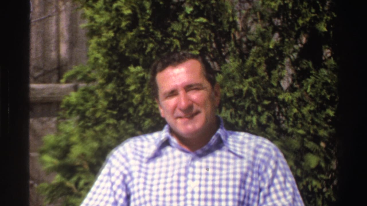 The man smiles toward the camera in this 1969 Michigan home-movie frame, seated before green shrubs in the yard.