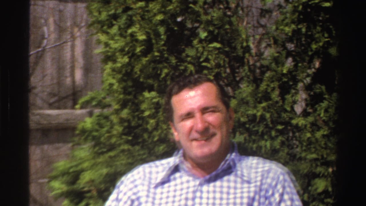 Man in a blue and white checked shirt smiles outdoors in front of leafy shrubs.