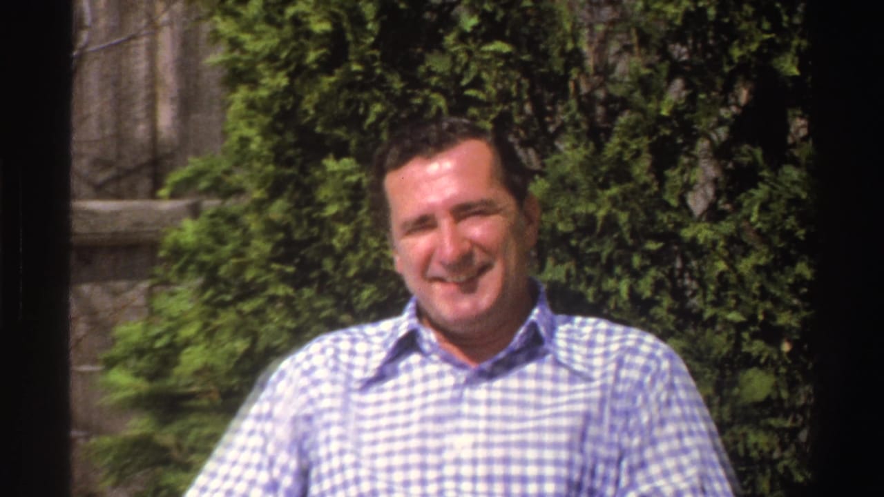 The man remains seated outdoors, smiling in bright sunlight with shrubs and a garden structure behind him.