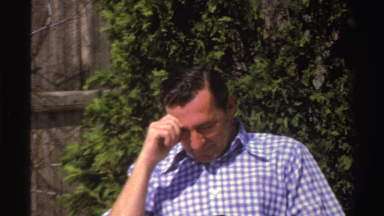 The seated man lowers his gaze and touches his head, with the book partly out of view and foliage behind him.