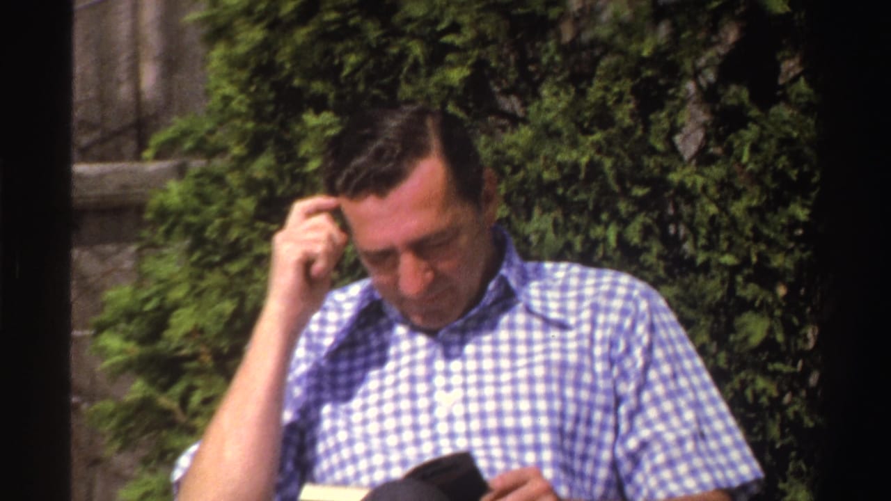 The man looks down toward a book in his lap while seated outside among greenery, matching the supplied 1969 Michigan home-movie context.