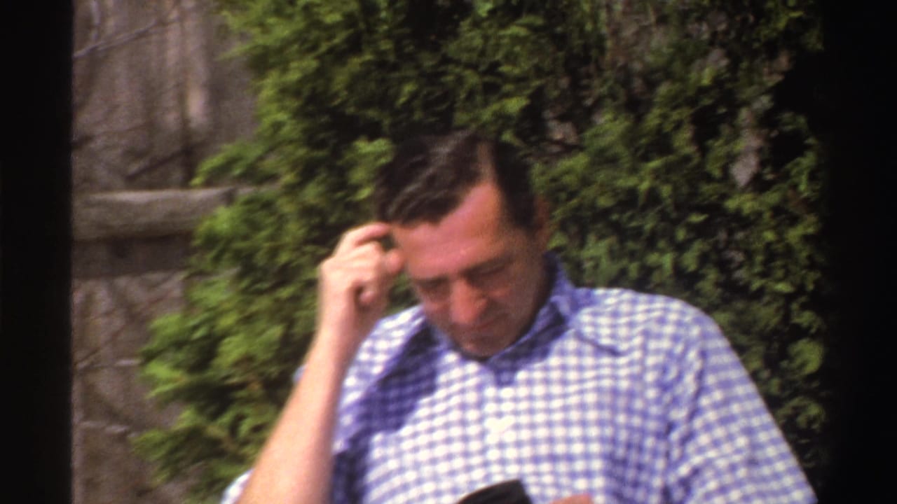 Man in a blue-and-white checkered shirt sits outdoors beside shrubs and a wooden fence.