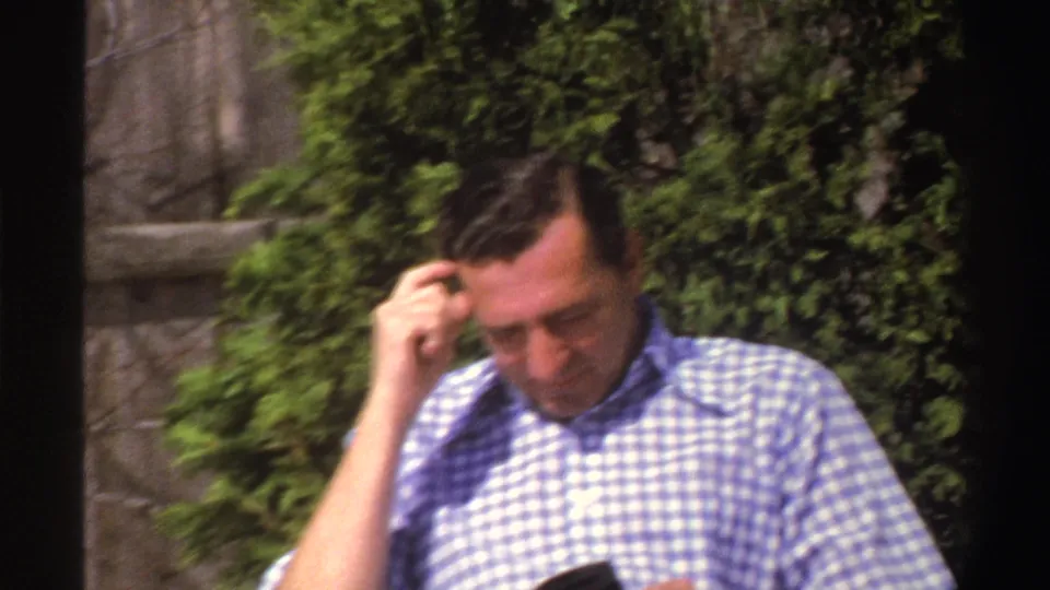 In 1969 Michigan home-movie footage, a man sits in a sunlit yard with his hand near his head, backed by evergreen shrubs and a wooden fence.
