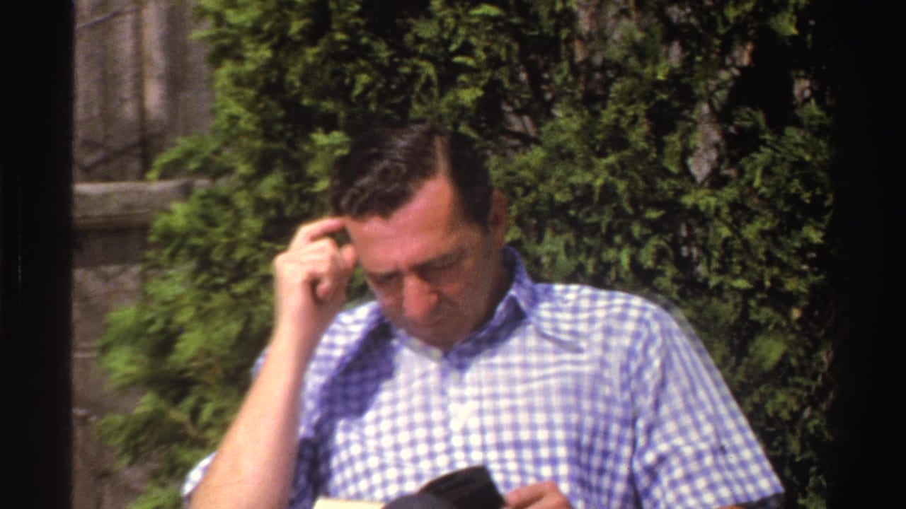 The man in a checkered shirt looks down at a small dark book or object while seated in a Michigan yard with shrubs and a wooden fence.