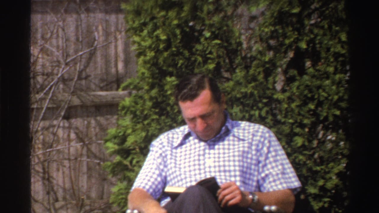 A wider 1969 Michigan frame shows the man seated outdoors, holding a small book or object with wooden fencing and greenery behind him.