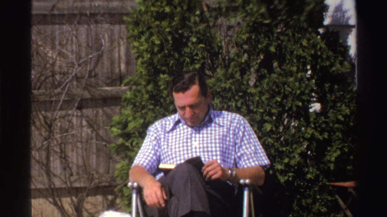 In a 1969 Michigan backyard, a man in a blue checked shirt reads in a folding chair near snow, shrubs, and a wooden fence.