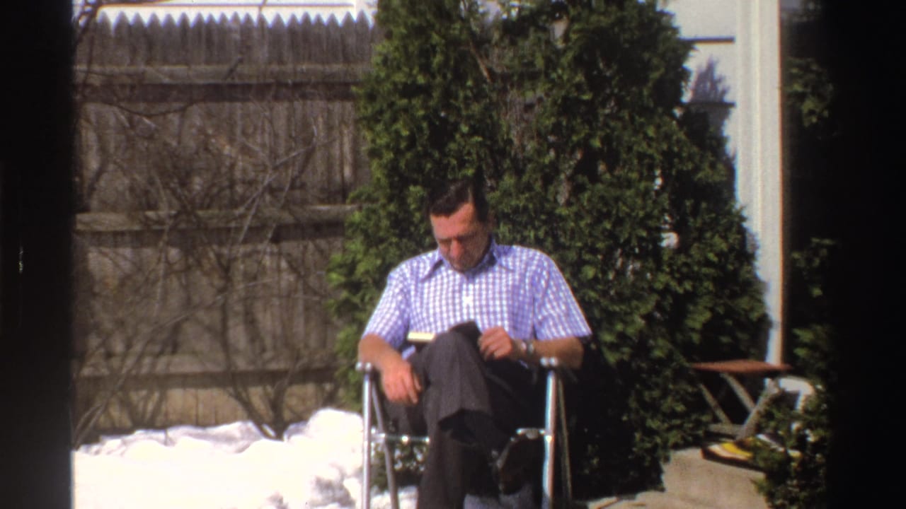 A man reads while seated in a folding chair in a Michigan backyard with remaining snow, evergreen shrubs, fence, and house siding visible.