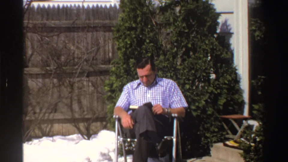 A man reads while seated in a folding chair in a Michigan backyard with remaining snow, evergreen shrubs, fence, and house siding visible.