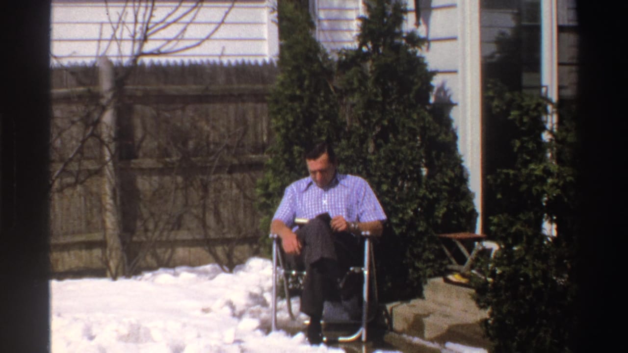 A wider 1969 Michigan home-movie frame shows the man reading in a folding chair, surrounded by snow, shrubs, fence, and residential siding.