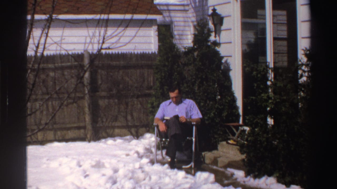 The man remains seated reading in the snowy Michigan backyard, with evergreen shrubs, fence, house trim, and porch area in view.