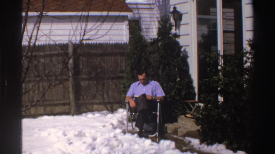 The man remains seated reading in the snowy Michigan backyard, with evergreen shrubs, fence, house trim, and porch area in view.