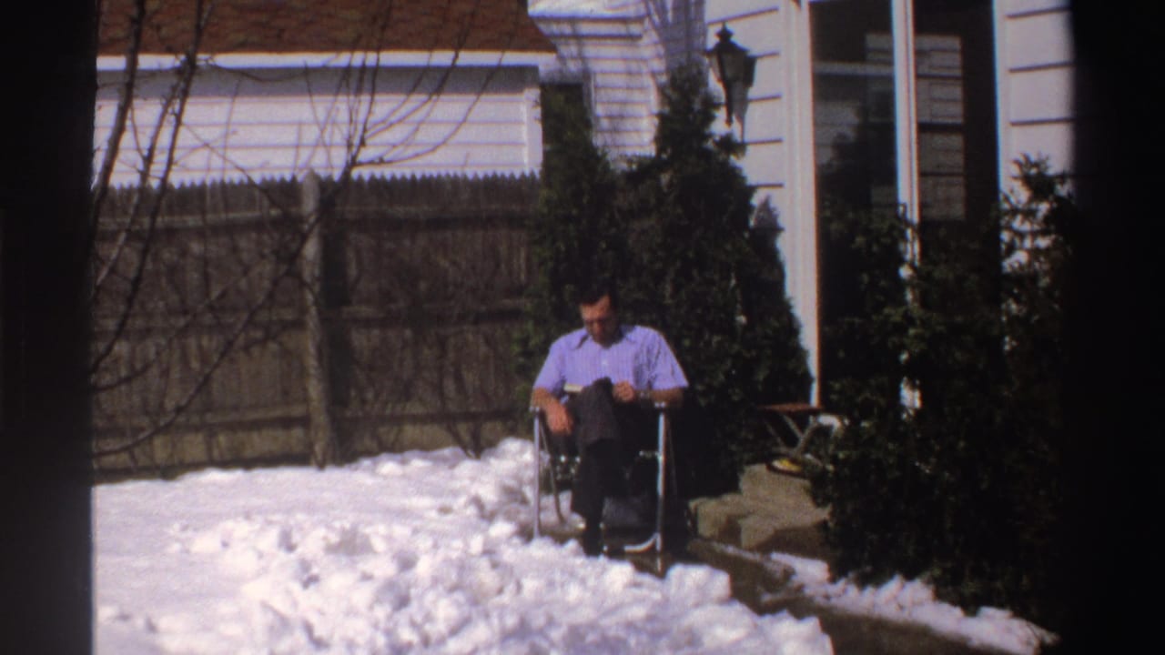 Man seated in a folding chair reading on a snowy patio beside a white house.