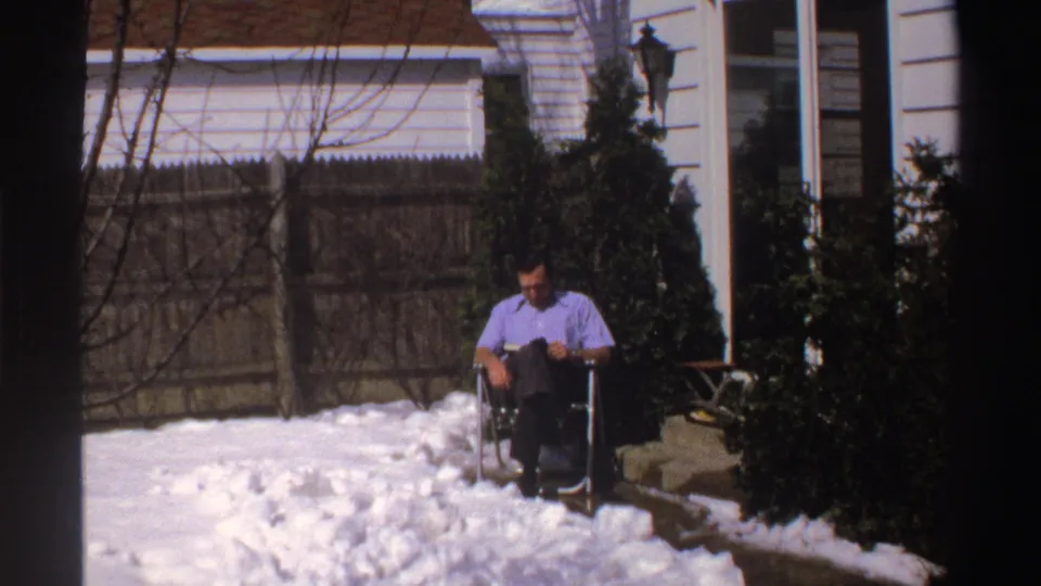 In Michigan 1969 home-movie footage, a man in a light purple shirt reads while seated on a snowy residential patio near shrubs and a wood fence.
