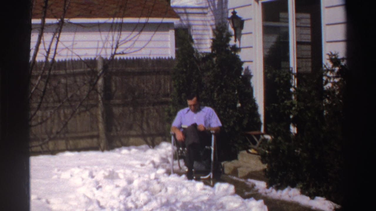 A man sits in a folding chair and looks down at reading material on a snowy Michigan patio, with a white house exterior and evergreen shrubs behind him.