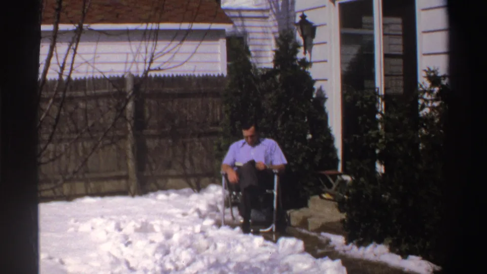 A man sits in a folding chair and looks down at reading material on a snowy Michigan patio, with a white house exterior and evergreen shrubs behind him.