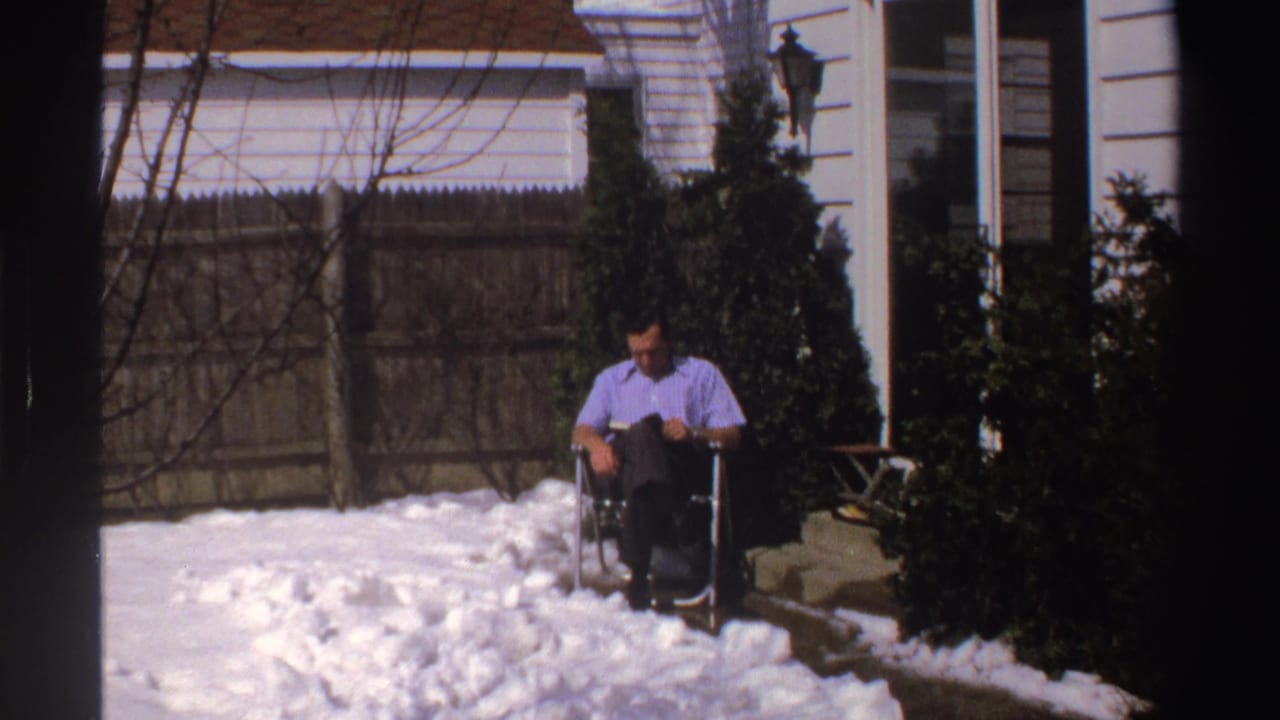 The home-movie frame shows the man reading in a folding chair, surrounded by snow, bare branches, a wooden fence, and the side of a white house.