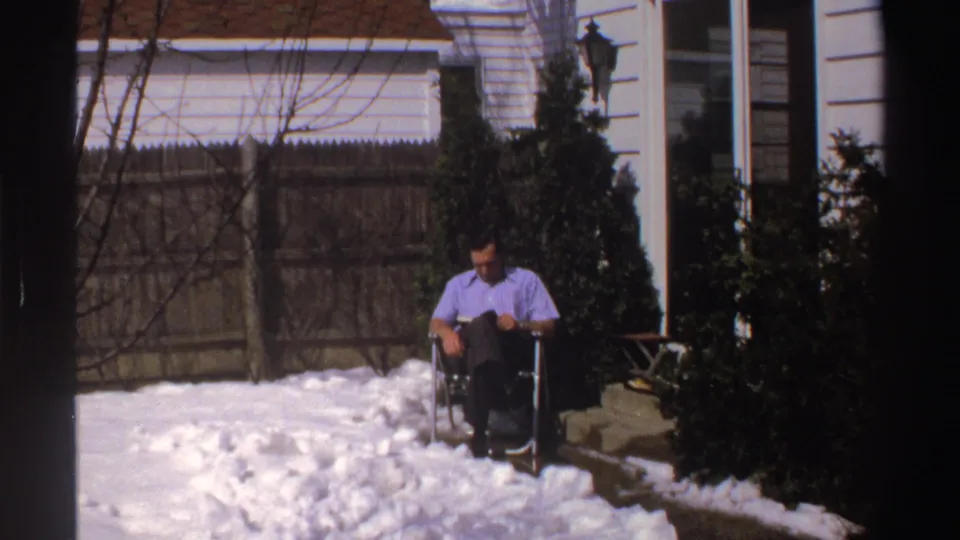 The home-movie frame shows the man reading in a folding chair, surrounded by snow, bare branches, a wooden fence, and the side of a white house.