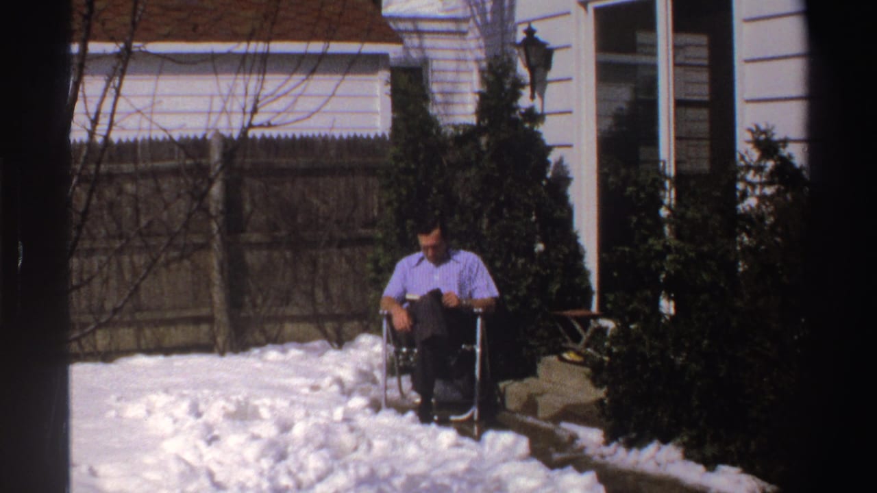 A man in a light purple button-down shirt reads on a snowy Michigan patio, framed by evergreen shrubs, a wood fence, and a white house exterior.