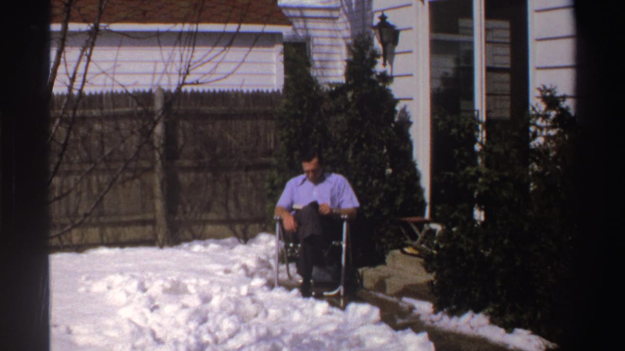 In 1969 Michigan home-movie footage, a man in a purple shirt sits in a folding chair in a snow-covered backyard near shrubs and house siding.