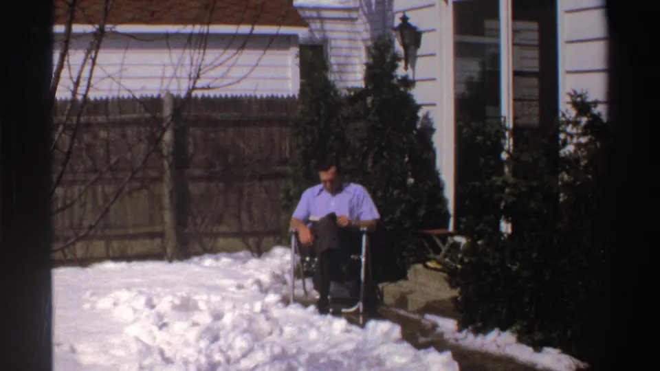 In 1969 Michigan home-movie footage, a man in a purple shirt sits in a folding chair in a snow-covered backyard near shrubs and house siding.