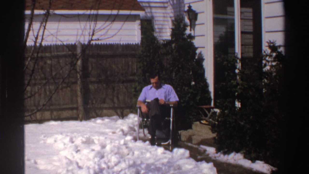 Man seated in snow near a fence, shrubs, and the side of a house.