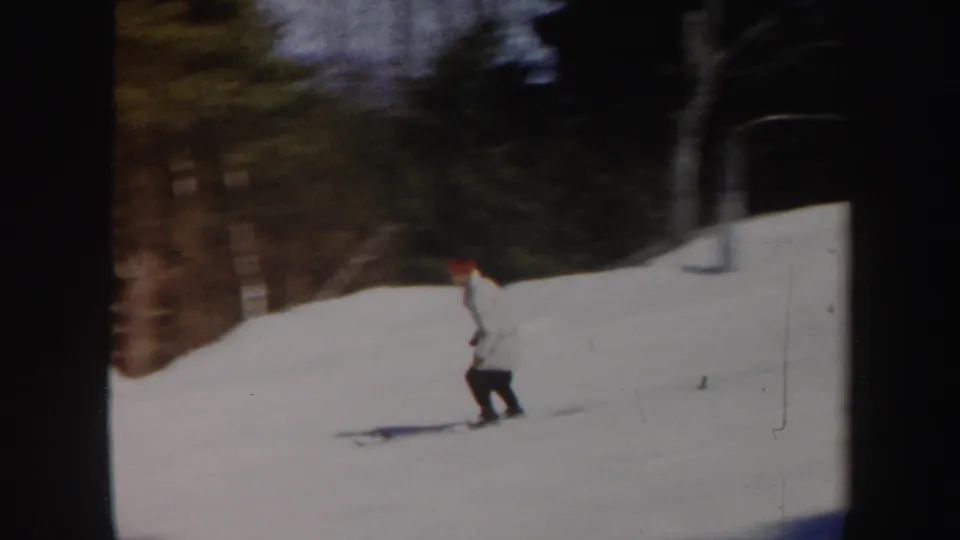The skier continues downhill on a snow-covered Vermont slope, with evergreens, bare trees, and dark film edges visible.