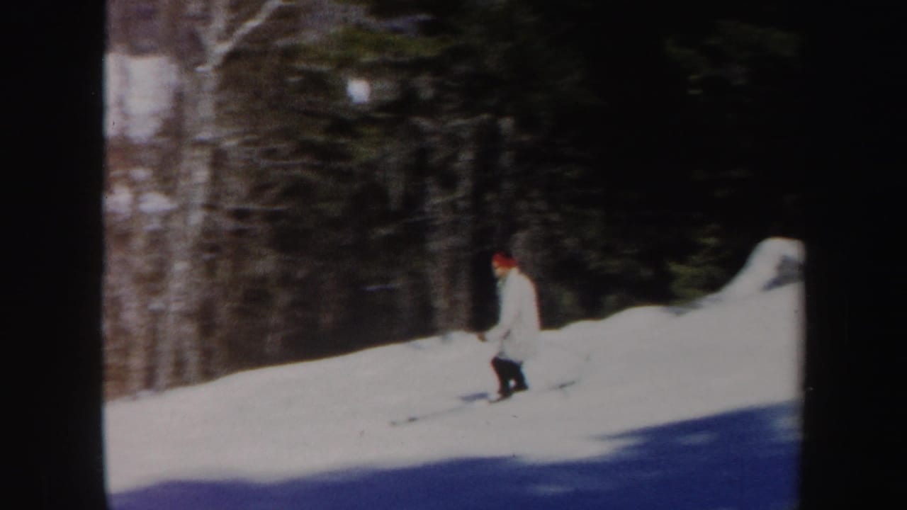 1962 Vermont home-movie frame showing a skier in a light coat and red hat on a snowy wooded slope.