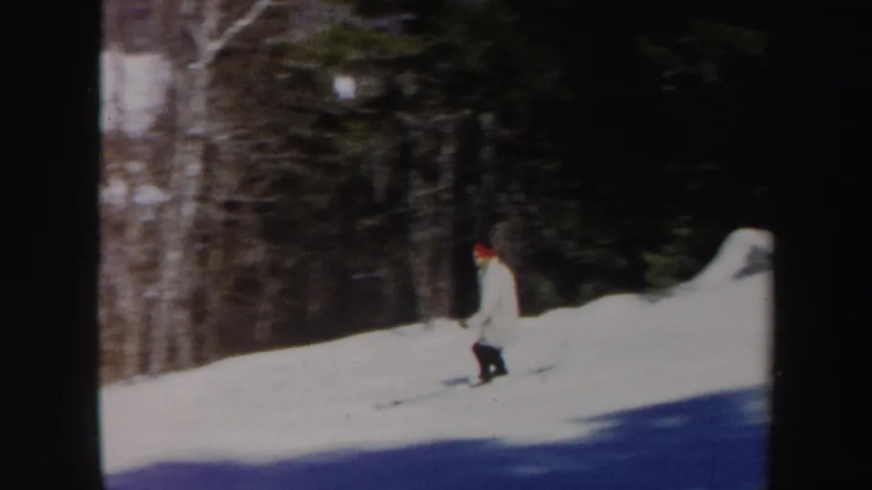 1962 Vermont home-movie frame showing a skier in a light coat and red hat on a snowy wooded slope.