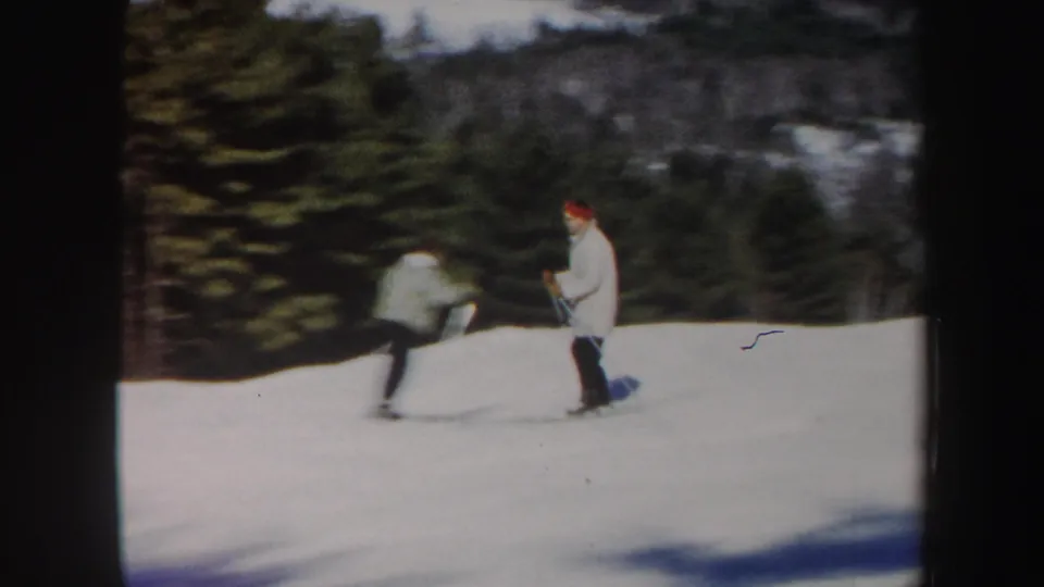 1962 Vermont frame with two skiers on a bright snowy slope bordered by evergreens and distant woods.