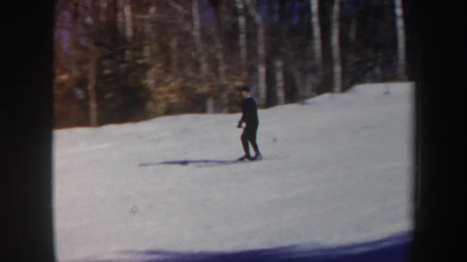 1962 Vermont home-movie frame showing a skier in dark clothing on an open snowfield below bare trees.