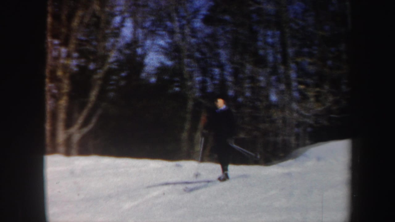 Man in a dark suit skis across a snowy slope near leafless trees.