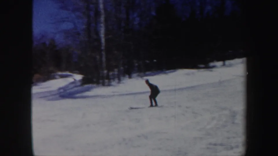1962 Vermont frame showing a lone skier in dark clothing descending a snowy slope with trees and long blue shadows behind them.