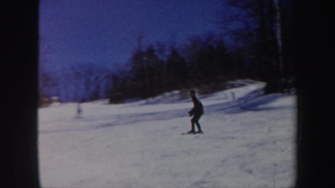 In 1962 Vermont 8mm footage, a distant skier in dark clothing moves across a snowy wooded slope under a blue sky.
