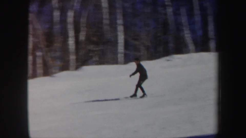 A dark-clad skier glides across a snow-covered slope near a line of trees in vintage 8mm home-movie footage.