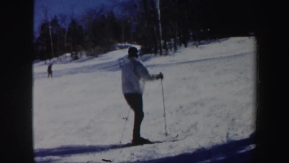 The skier appears to slow or pause on the Vermont ski slope, with bare trees and another skier in the background.