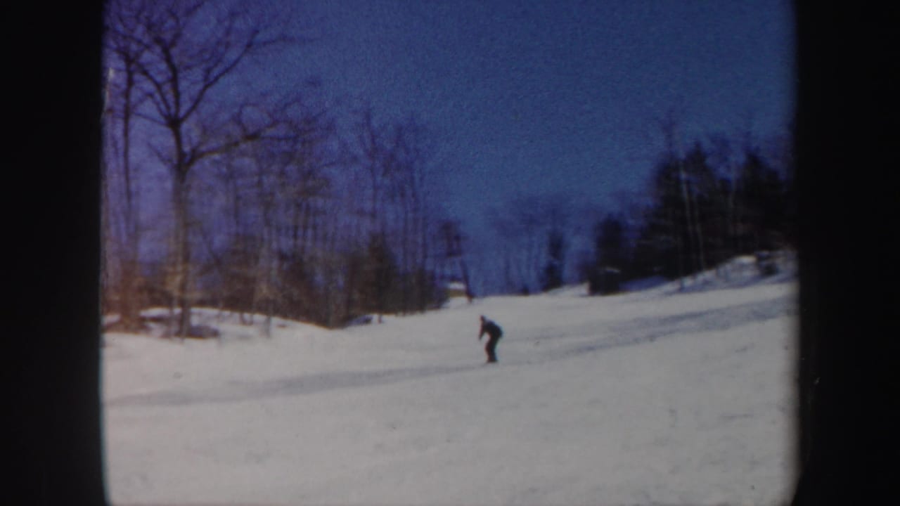A distant skier in dark clothing crosses a snowy Vermont slope in 1962 home-movie footage, with bare trees and blue sky behind.