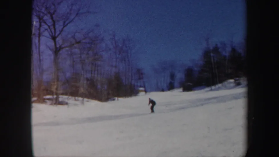 A distant skier in dark clothing crosses a snowy Vermont slope in 1962 home-movie footage, with bare trees and blue sky behind.