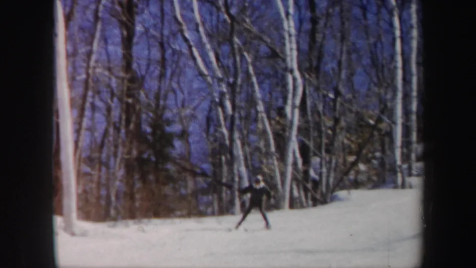 A dark-clad skier glides across the snow in 1962 Vermont footage, with tall white-barked trees filling the background.