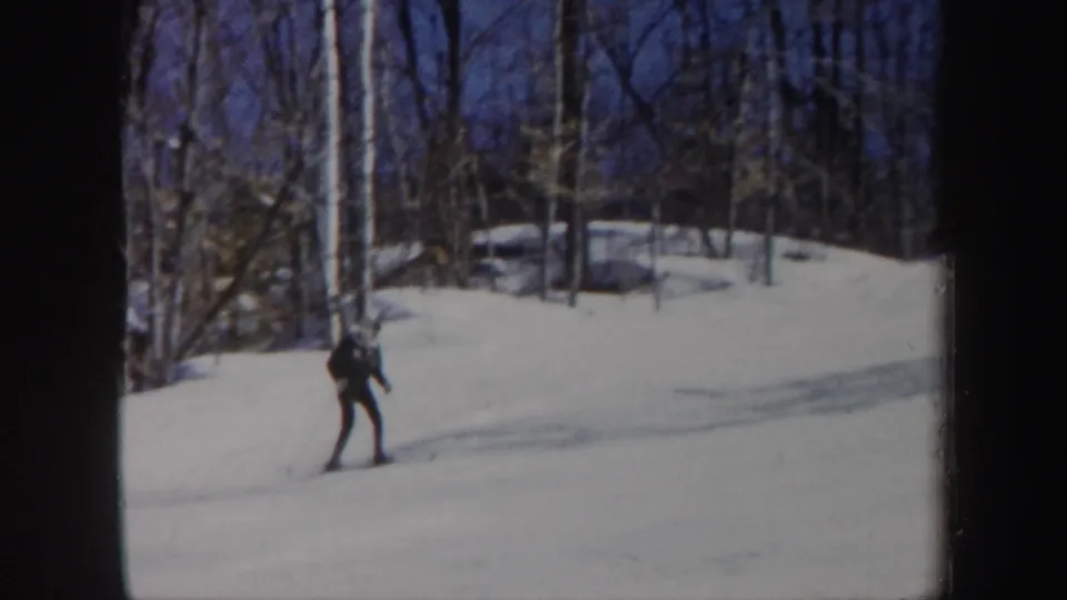 A skier in dark winter clothing moves across a snow-covered Vermont slope in 1962 home-movie footage.