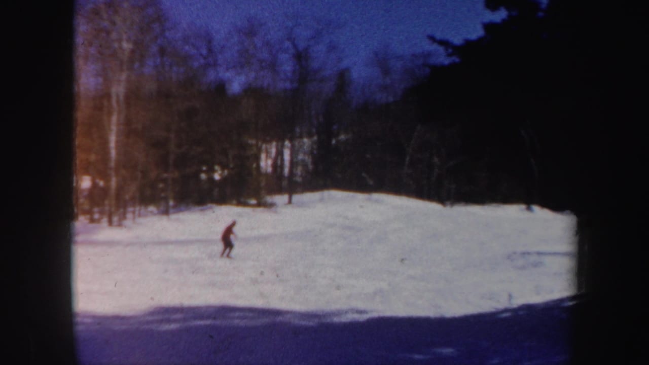 Distant skier in a red jacket crosses a snowy slope bordered by trees.