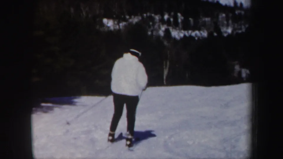 A second adult skier in a white jacket is seen from behind on the snowy Vermont hill, holding ski poles near the tree line.