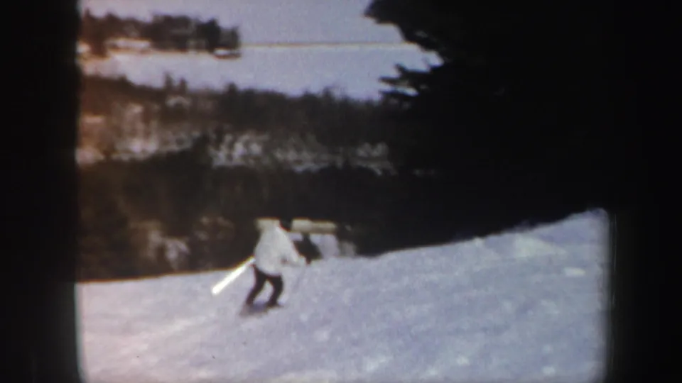 1962 Vermont 8mm frame showing a lone skier moving downhill across snow, with dark forest and a distant pale body of water in the background.