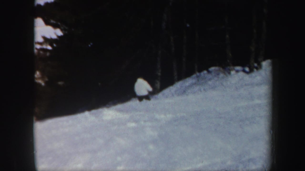 1962 Vermont home-movie frame with a distant skier crossing a snow-covered hillside near a dark evergreen tree line.